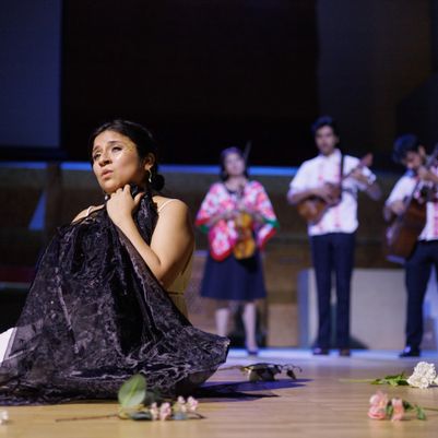 Mariela sits on a stage clutching a black cloth, as musicians in colorful attire play behind her, with flowers scattered on the floor