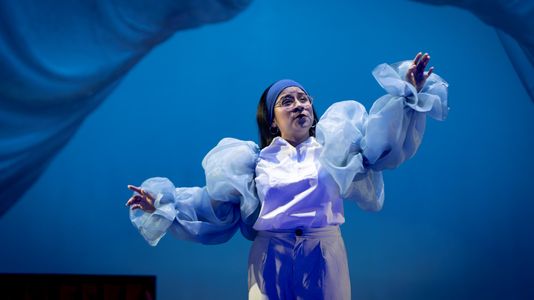 Mariela on stage gestures expressively, wearing a headband and flowing sheer sleeves. The blue backdrop creates a dramatic ambiance.