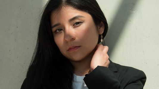 Mariela stands against a light wall, wearing a black blazer and white shirt. She gazes thoughtfully, holding her neck.