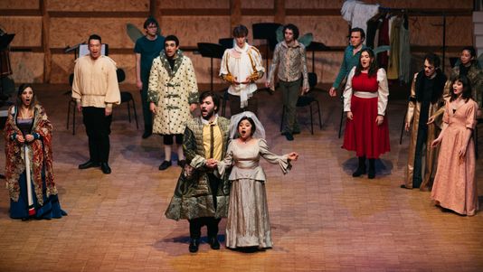 Mariela and other performers in vivid medieval costumes sing expressively on a wooden stage