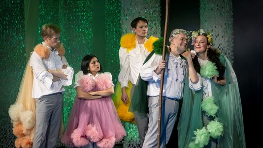 Mariela and four other actors in colorful, whimsical costumes perform on stage. One actor holds a staff, smiling with another wearing a floral crown, creating a joyful mood.