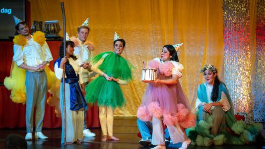 A joyful group of actors in colorful costumes with party hats stands on stage against a golden backdrop. Mariela holds a cake, celebrating with smiles and laughter.